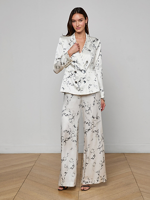 A woman stands indoors on a wooden floor wearing the Colin Silk Blazer and matching white pants with a black abstract print, paired with open-toe black heels. She faces forward relaxedly against a plain light-colored wall.