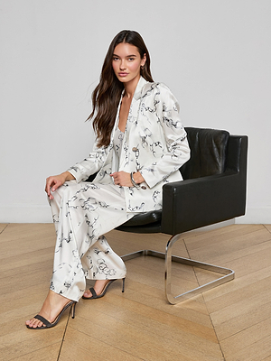 A woman with long brown hair sits on a modern black armchair, wearing the Colin Silk Blazer with a black abstract pattern and high-heeled sandals, on a light wood floor against a plain white wall.