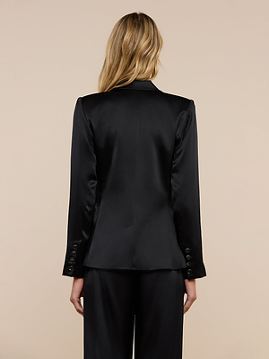 A woman with long, wavy blonde hair is shown from the back wearing the Colin Silk Charmeuse Blazer, paired with matching trousers, against a plain beige background.