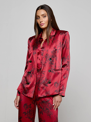 A woman with long straight hair wears the L'AGENCE Colin Silk Blazer—burgundy, shiny, and floral—with matching pants, standing against a plain light gray background. She looks confidently at the camera with a neutral expression.