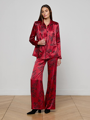 A woman poses confidently against a light gray wall, wearing the L'AGENCE Colin Silk Blazer—a shiny double-breasted pantsuit with a subtle black floral pattern—paired with black heels. She has long hair and one hand on her blazer.