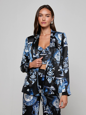 A woman with long brown hair wears the L'AGENCE Colin Silk Blazer, part of a blue and white porcelain print three-piece suit, featuring a matching crop top and high-waisted pants, against a plain light gray background.