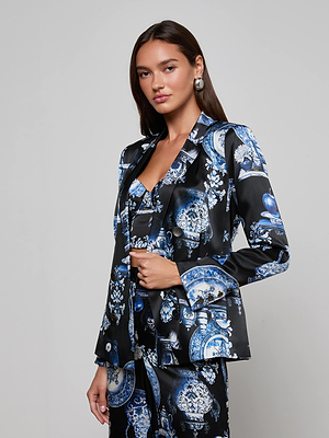 A woman with long brown hair models the L'AGENCE Colin Silk Blazer, a black silk charmeuse suit adorned with blue and white porcelain print. She stands against a light gray background, gazing confidently at the camera with one hand tucked in her jacket.