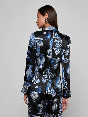 A woman with long brown hair faces away, modeling the L'AGENCE Colin Silk Blazer—a blue and white porcelain print double-breasted set in black silk charmeuse—against a plain light gray wall.