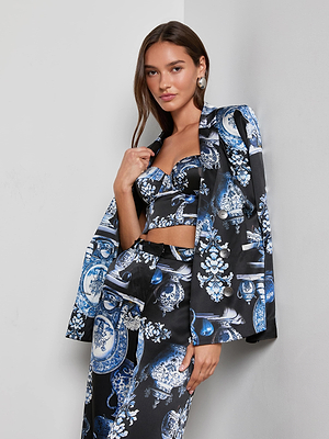 A woman with long brown hair and large silver earrings stands against a white wall wearing the L'AGENCE Colin Silk Blazer, draped over her shoulders, paired with a matching crop top and high-waisted pants in a blue and black porcelain print.
