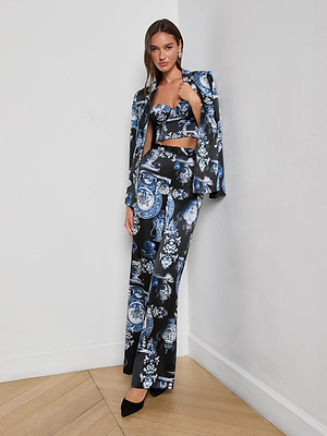 A woman stands in a corner wearing Pilar Silk Wide-Leg Pants as part of a blue and white porcelain print three-piece set with a blazer and crop top. She pairs the look with black pointed heels and has long, straight hair.