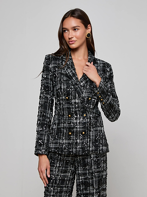 A woman wears the L'AGENCE Colin Sequined Tweed Blazer, a black and white plaid double-breasted style with gold buttons, standing against a light gray background and looking slightly to the side while touching her collar.