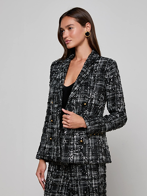 A woman with long brown hair wears the L'AGENCE Colin Sequined Tweed Blazer with gold buttons and matching pants, paired with a black top and round black earrings, standing against a light gray background.