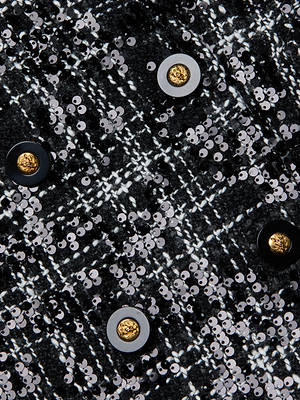 A close-up of the L'AGENCE Colin Sequined Tweed Blazer reveals black and white fabric with shiny silver sequins, round black and gold buttons, and intersecting white lines for a sophisticated tailored look.