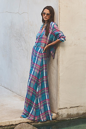 A woman stands by a light-colored wall, wearing sunglasses and a long, colorful plaid dress with long sleeves. She has long hair and is standing near the edge of a small pool of water.