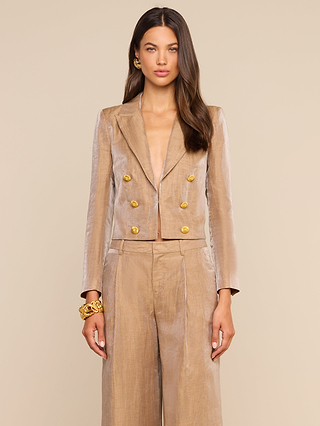 A woman stands facing forward, wearing the Inez Cropped Blazer—a shiny gold double-breasted design with gold buttons—paired with matching wide-leg pants. Her hands rest by her sides as she showcases long, wavy brown hair, gold earrings, and a chunky gold bracelet.