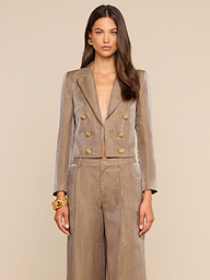 A woman stands against a white background wearing the Inez Cropped Blazer, a taupe linen blend double-breasted style with gold buttons, paired with matching wide-leg pants. She has long, wavy brown hair and completes her look with gold hoop earrings and a chunky gold bracelet.