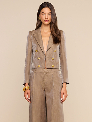 A woman faces forward, wearing the Inez Cropped Blazer in shiny taupe as part of a pantsuit featuring gold buttons, accessorized with a gold bracelet and earrings. She has long brown hair styled down, neutral makeup, and is posed against a beige background.