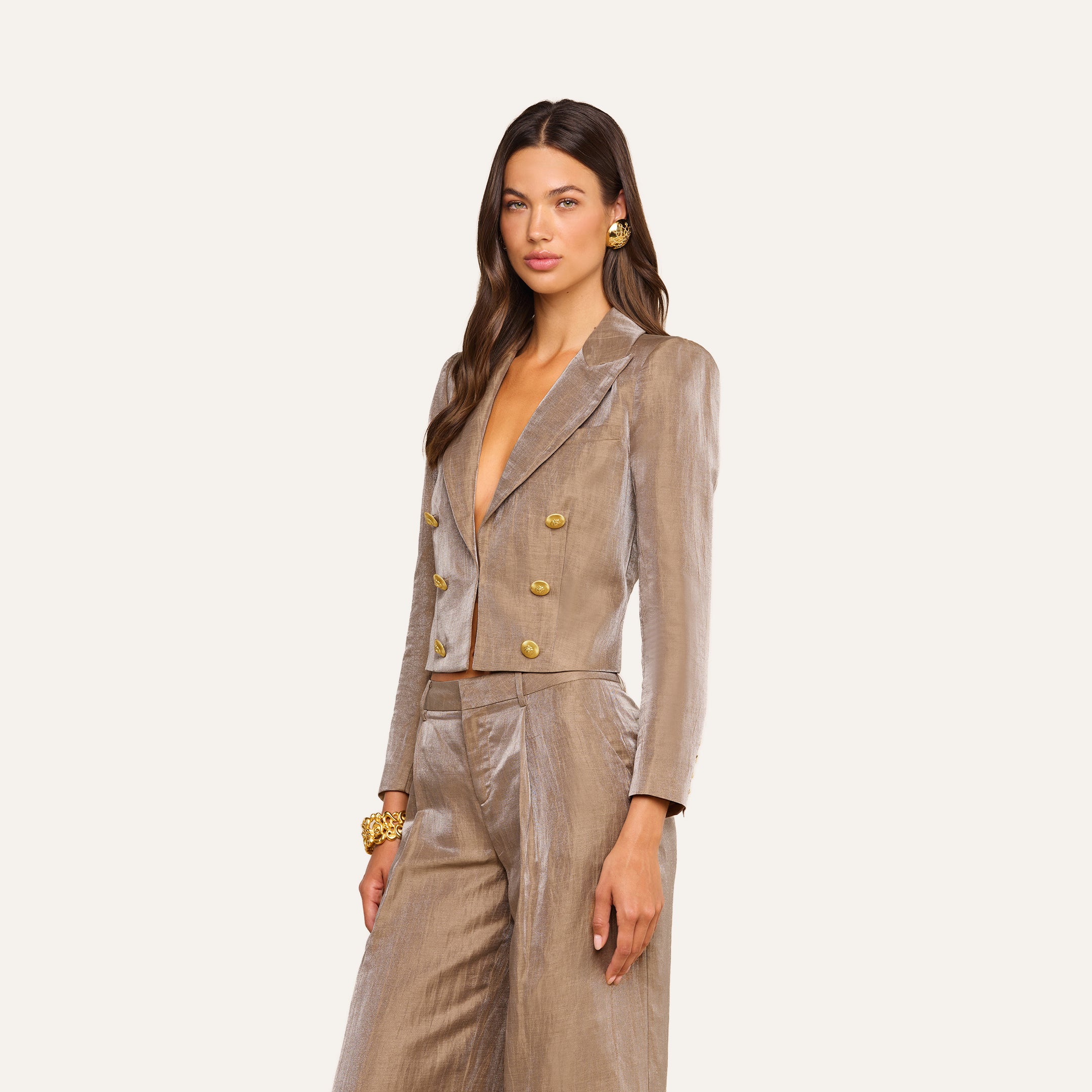 A woman poses in the Inez Cropped Blazer, a light brown, double-breasted linen blend suit jacket with gold buttons. She has long brown hair, gold earrings, and a bracelet. The background is plain and light-colored.