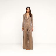 Une femme se tient debout devant un fond beige, vêtue du blazer court Inez — une veste en lin beige clair brillante et ajustée, ornée de boutons dorés —, quelle porte avec un pantalon assorti. Elle a de longs cheveux bruns, porte des bracelets dorés et des chaussures à bout pointu beiges, et regarde droit devant elle avec une expression neutre.