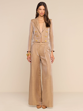 A woman stands against a beige background wearing the Inez Cropped Blazer—a shiny, tailored tan linen jacket with gold buttons—paired with matching pants. She has long brown hair, gold bracelets, and tan pointed-toe shoes, gazing forward with a neutral expression.
