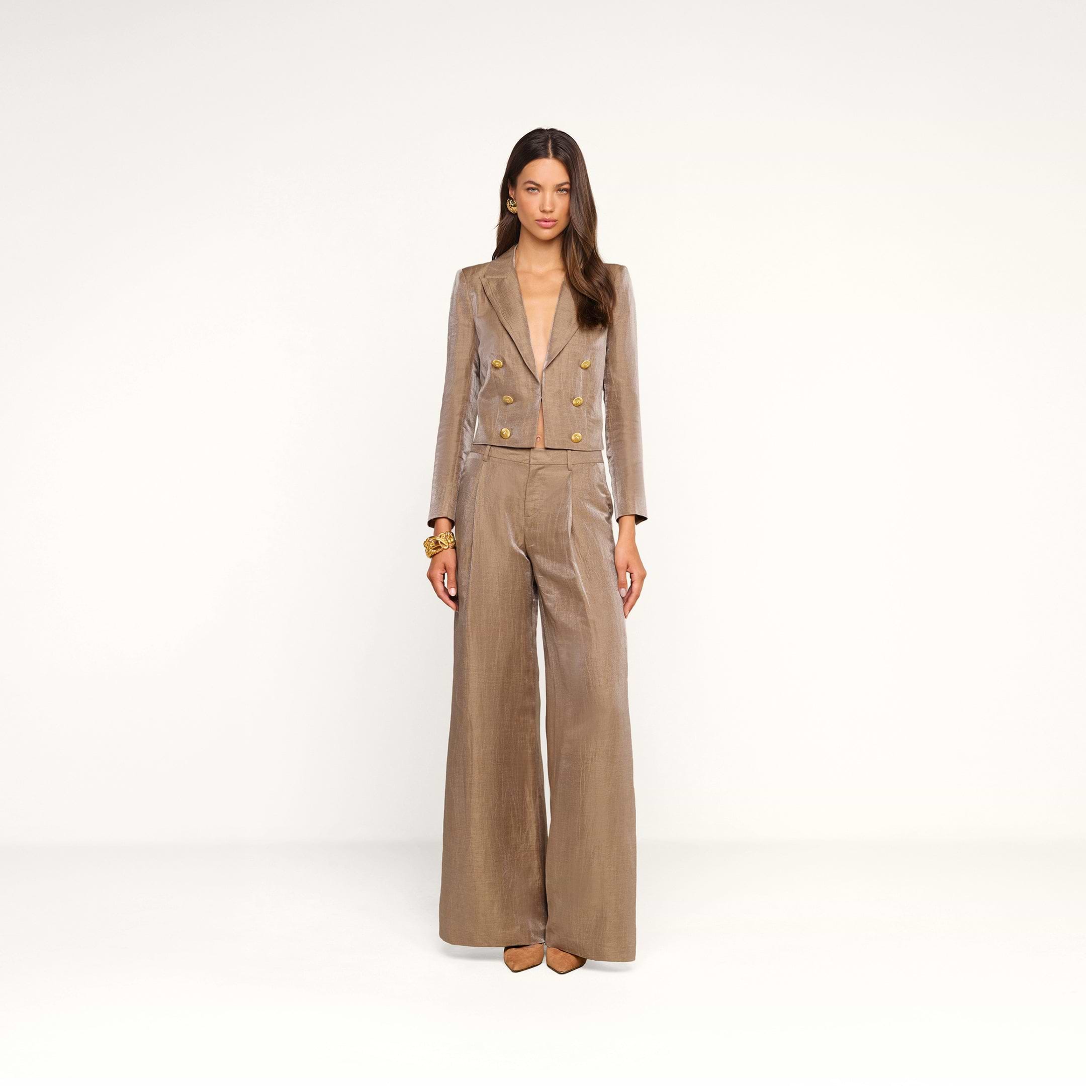Une femme se tient debout devant un fond beige, vêtue du blazer court Inez — une veste en lin beige clair brillante et ajustée, ornée de boutons dorés —, quelle porte avec un pantalon assorti. Elle a de longs cheveux bruns, porte des bracelets dorés et des chaussures à bout pointu beiges, et regarde droit devant elle avec une expression neutre.