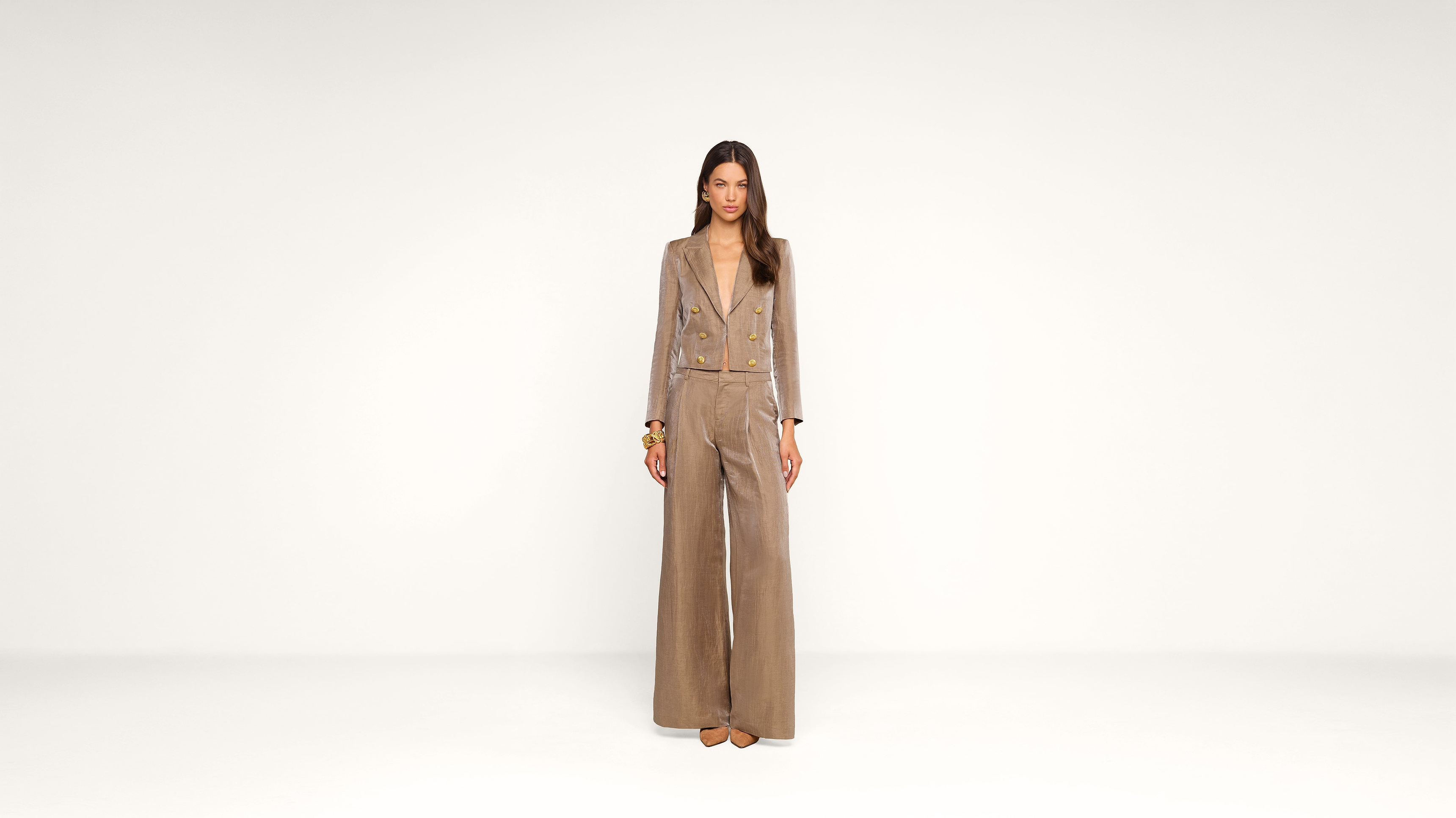 A woman stands in the center of a plain white background, wearing a tan, wide-legged pantsuit with gold buttons and beige heels. She has long, straight dark hair and a neutral expression.