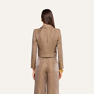 On voit de dos une femme aux longs cheveux châtains, debout devant un fond beige, vêtue du blazer court Inez — une veste en lin beige clair brillant ornée de boutons dorés.