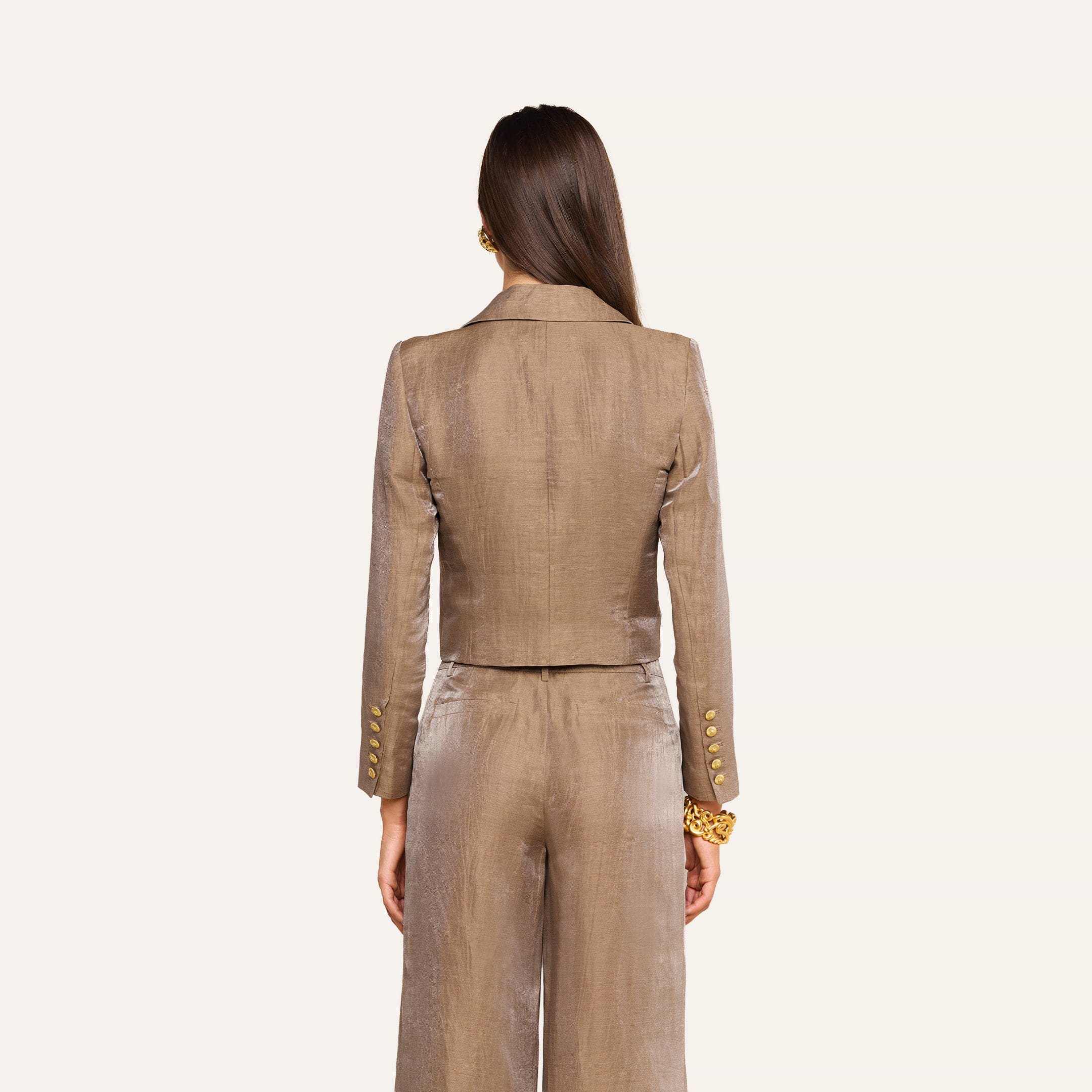 On voit de dos une femme aux longs cheveux châtains, debout devant un fond beige, vêtue du blazer court Inez — une veste en lin beige clair brillant ornée de boutons dorés.