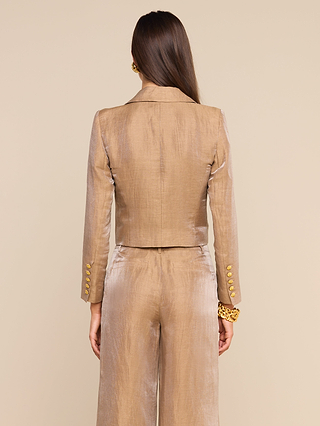 A woman with long brown hair is seen from the back, standing against a beige background, wearing the Inez Cropped Blazer—a shiny tan linen jacket with gold buttons.