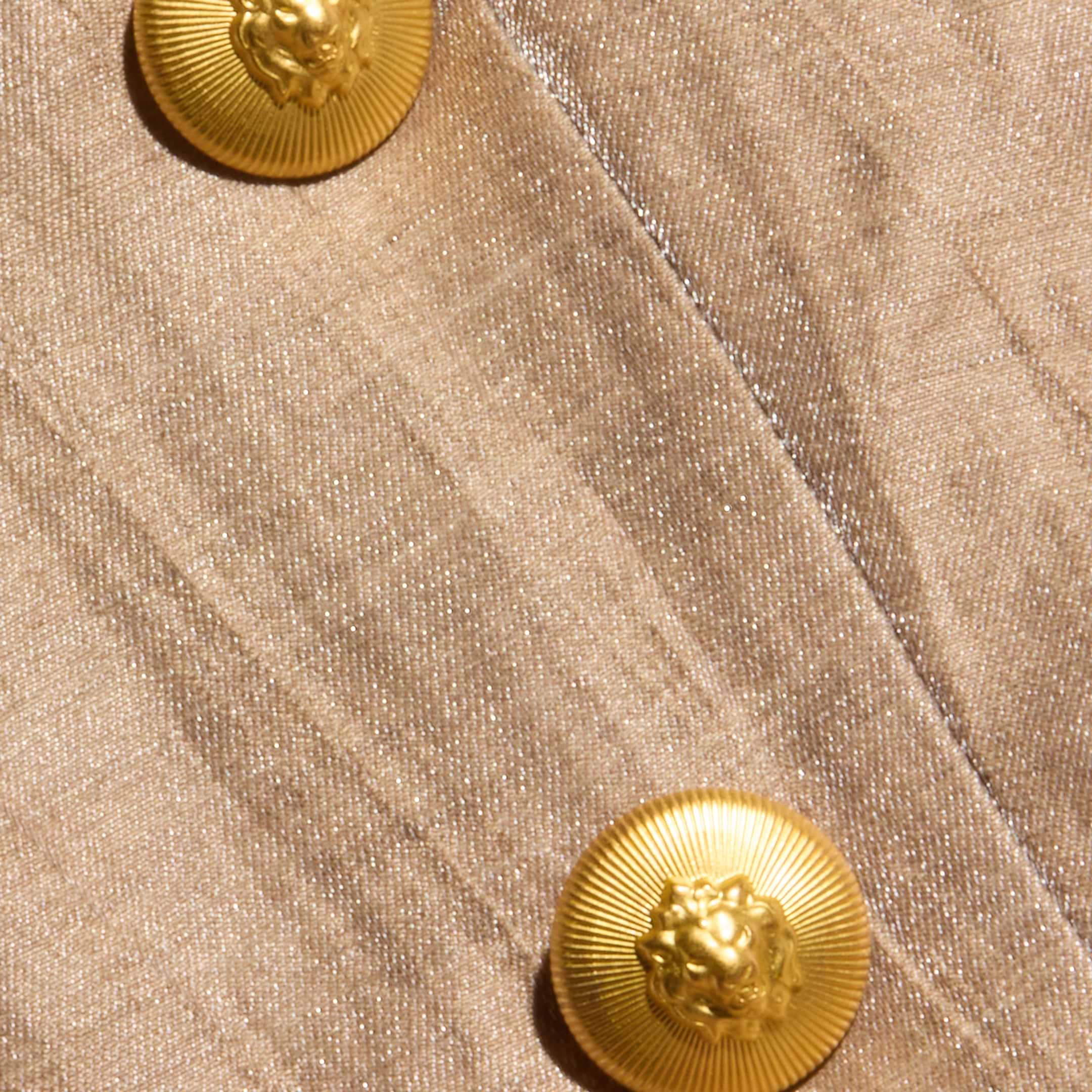 Close-up of the Inez Cropped Blazer’s beige linen blend fabric, detailed with two shiny, round gold buttons adorned with a raised lion’s head design. The fabric has a slightly textured, woven appearance.