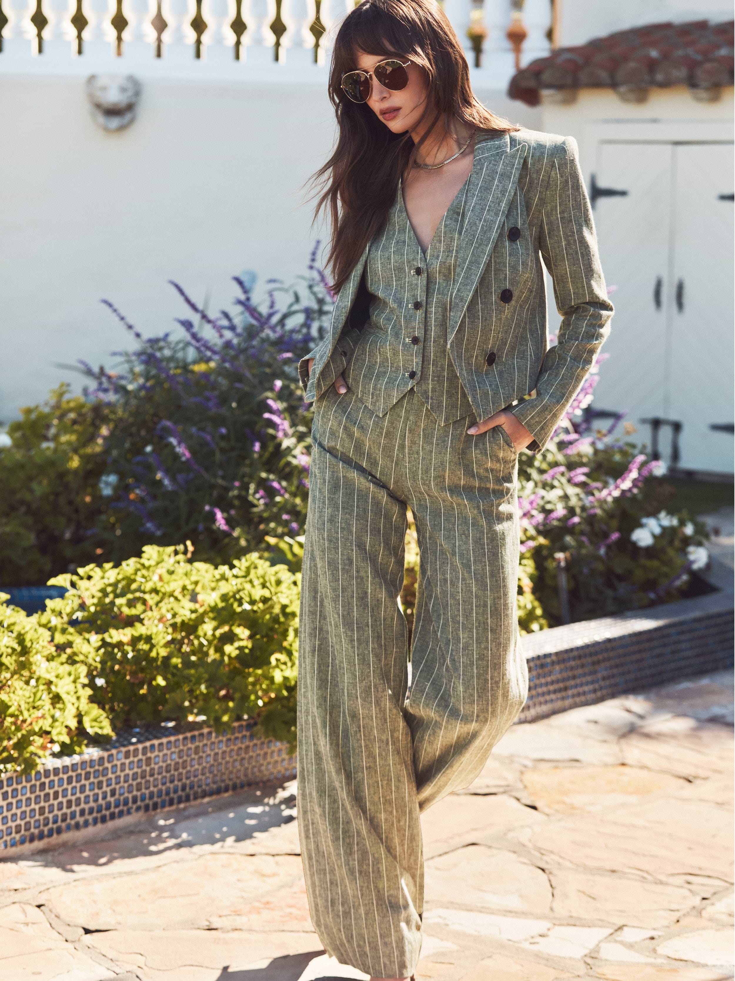 Woman wearing a light green pinstripe suit with wide-leg pants and a matching vest, standing outdoors in sunlight with one hand in her pocket, sunglasses on, and plants in the background.