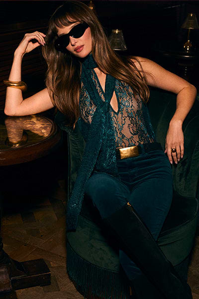A woman in dark sunglasses sits on a green velvet chair, wearing a sheer lace top, a scarf, dark velvet pants with a gold belt, and knee-high black boots. She has long brown hair with bangs and a gold bracelet.