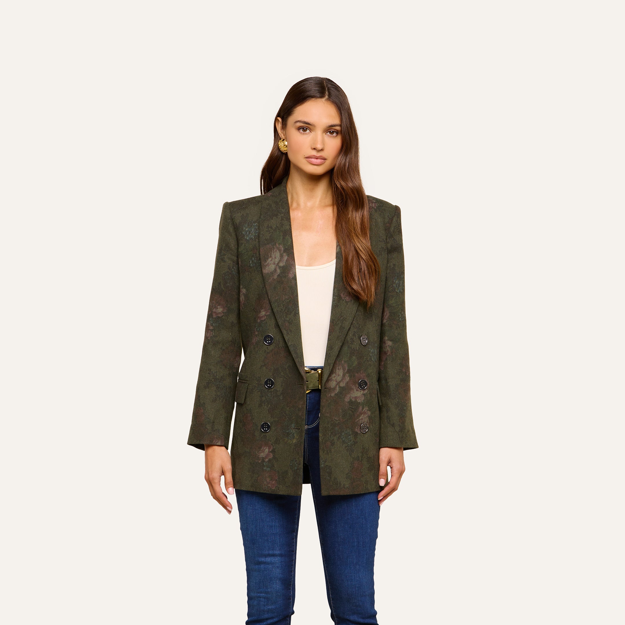 A woman stands facing forward, wearing the Jayda Blazer—a green floral print blazer with a double-breasted design—paired with blue jeans and a white top, against a plain light background.