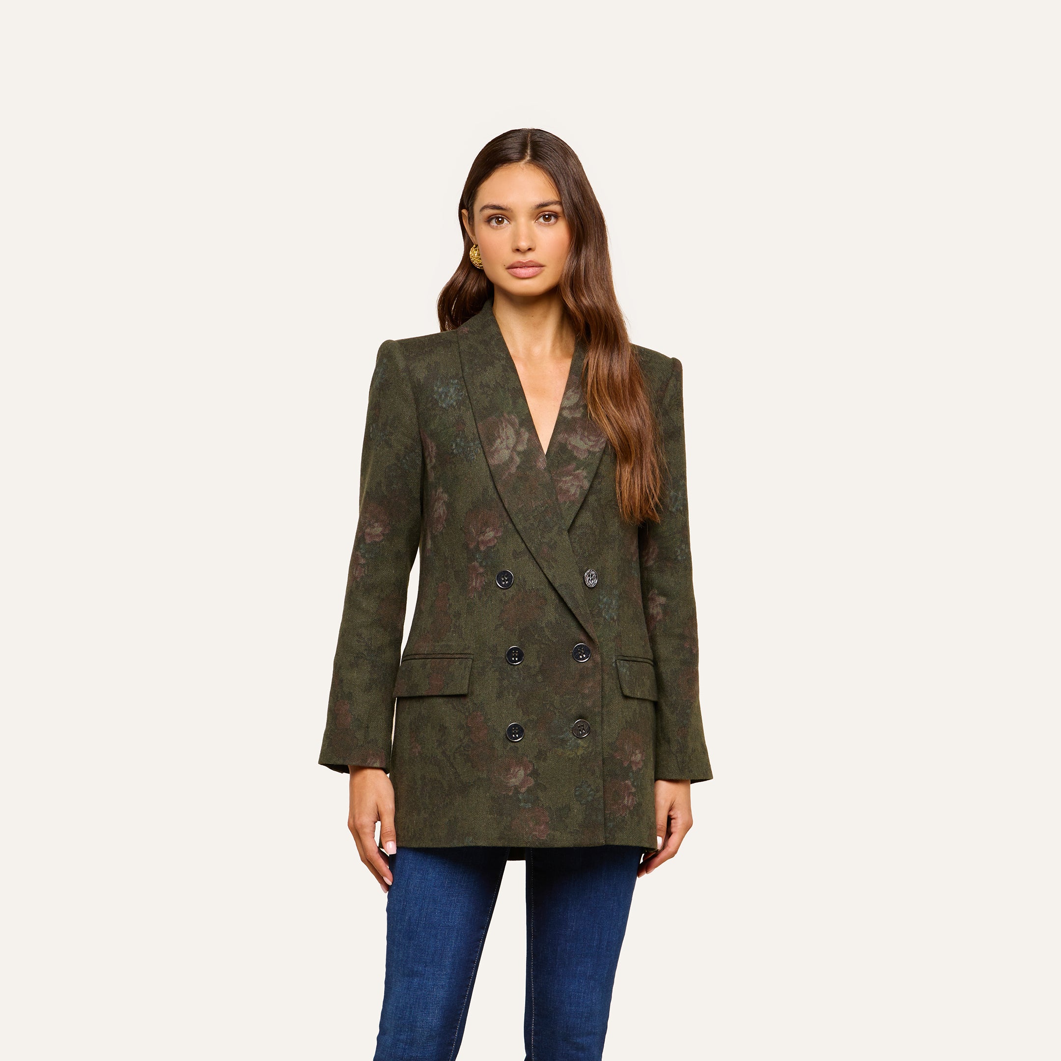 A woman with long brown hair wears the Jayda Blazer, a dark green floral print double-breasted piece, paired with blue jeans and standing against a plain light background.
