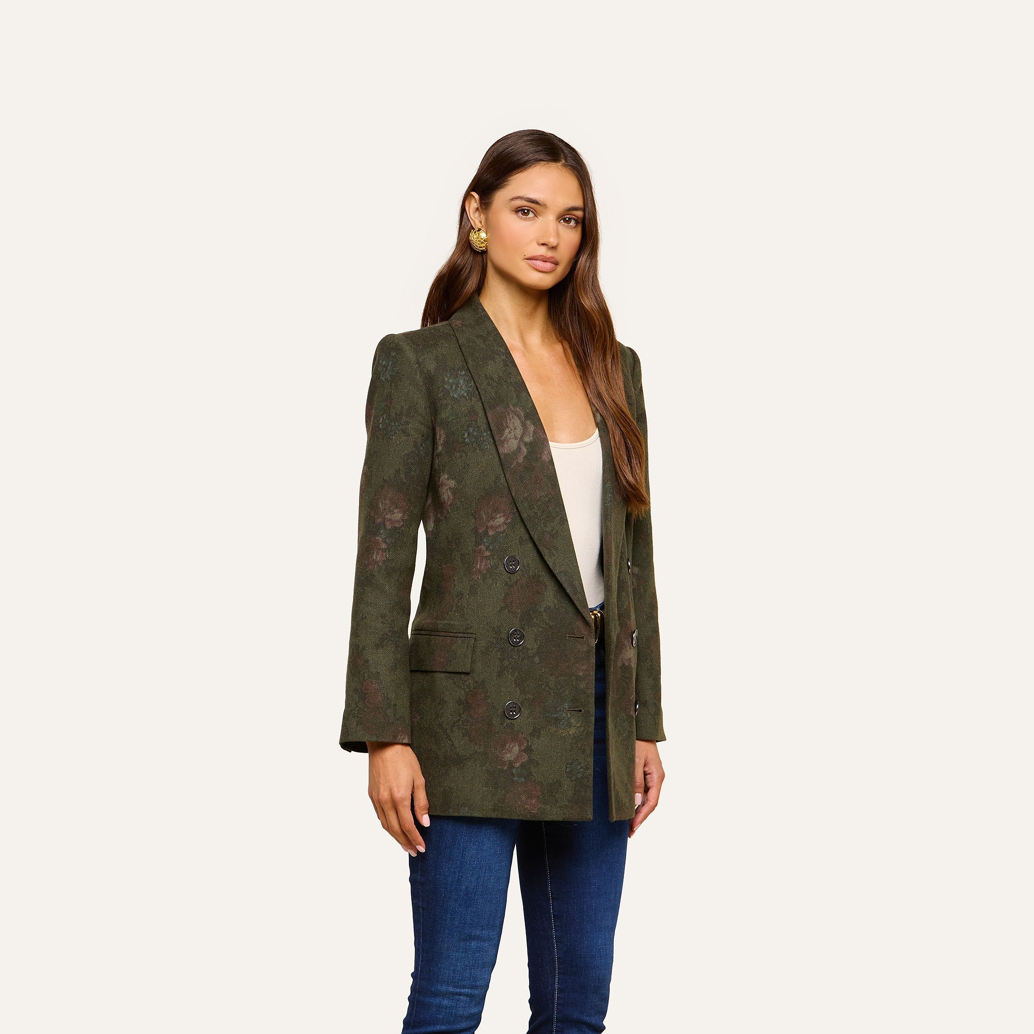 A woman wears the Jayda Blazer, featuring an oversized dark green floral print, layered over a white top and blue jeans. She stands against a plain light background, with long brown hair and gold earrings accenting her look.