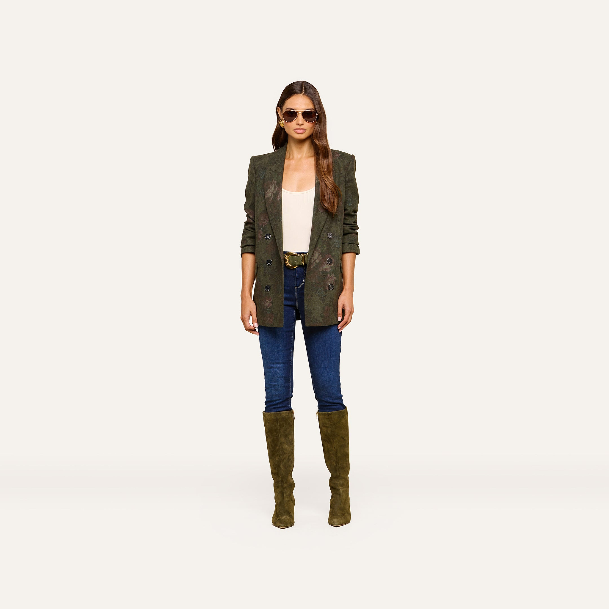 A woman wearing sunglasses, the Jayda Blazer in oversized olive green, a white top, blue jeans, and olive knee-high boots stands facing forward against a plain light background.