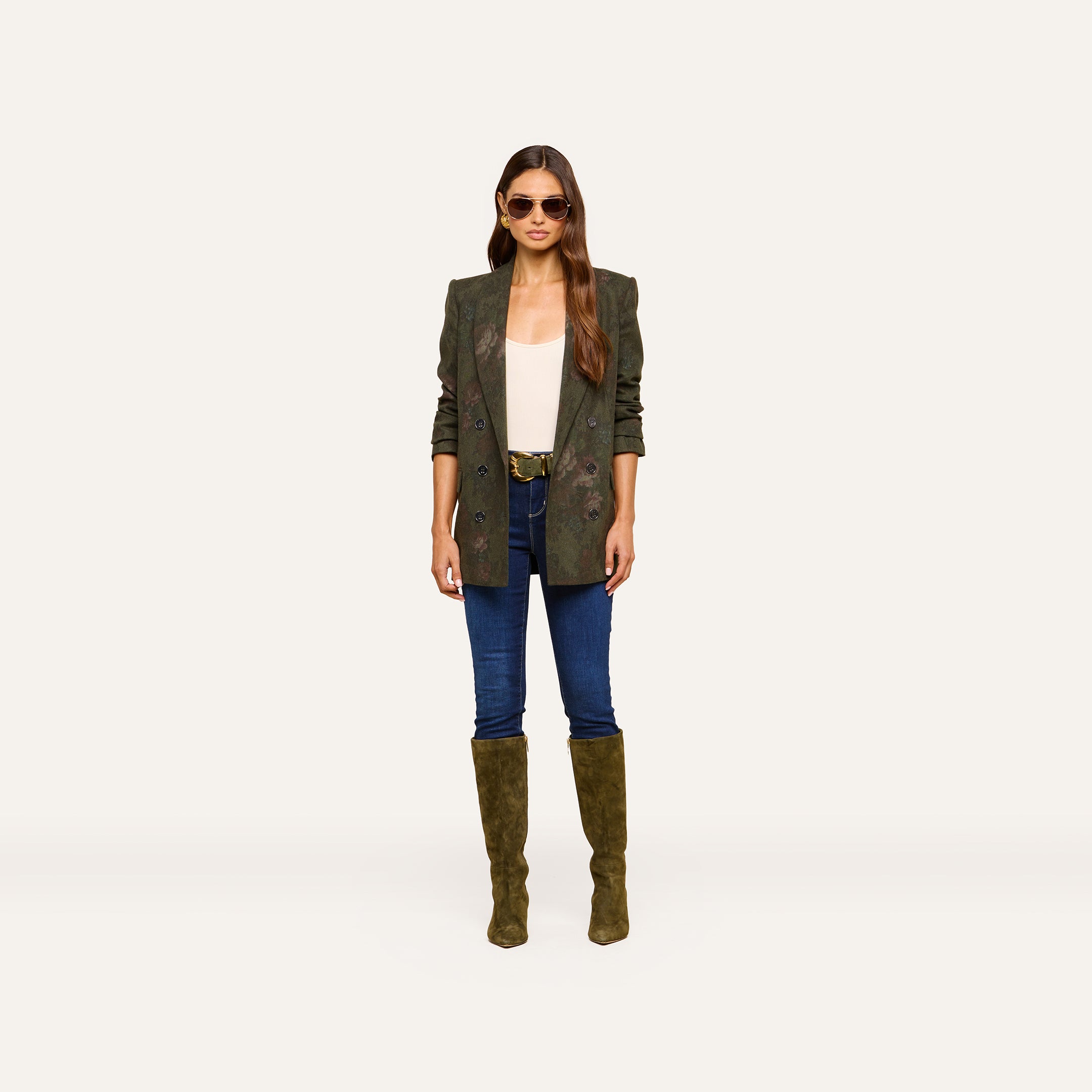 A woman wearing sunglasses, the Jayda Blazer in oversized olive green, a white top, blue jeans, and olive knee-high boots stands facing forward against a plain light background.