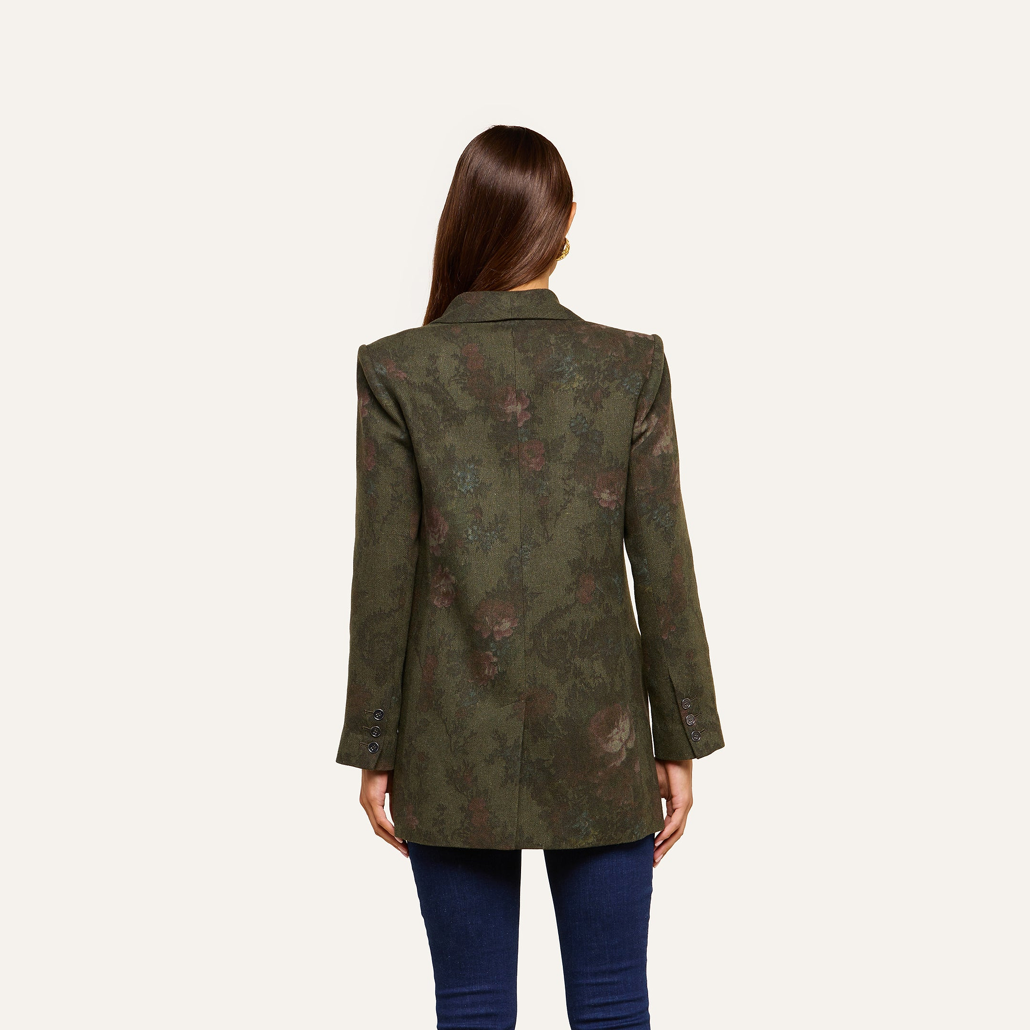 A woman with long brown hair faces away, wearing the Jayda Blazer—a dark green, floral print blazer—paired with dark blue jeans against a plain light background.