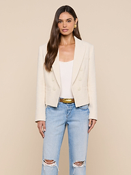 A woman stands facing forward against a plain light background, wearing the cream Brooke Tweed Blazer over a white top, paired with light blue ripped jeans and a gold belt.