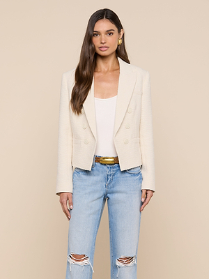 A woman wearing the Brooke Tweed Blazer in ecru over a white top and light blue ripped jeans stands against a beige background, her long brown hair styled down, accessorized with gold earrings and a neutral expression.