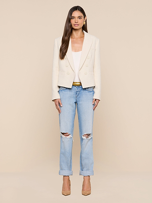 Woman with long brown hair and a neutral expression stands against a beige background, wearing the Brooke Tweed Blazer in ecru over a white top, paired with light blue ripped jeans and tan heels.