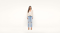 A woman stands against a plain white background, wearing a beige blazer, white top, ripped light blue jeans, and beige heels. She has long brown hair and a neutral expression.