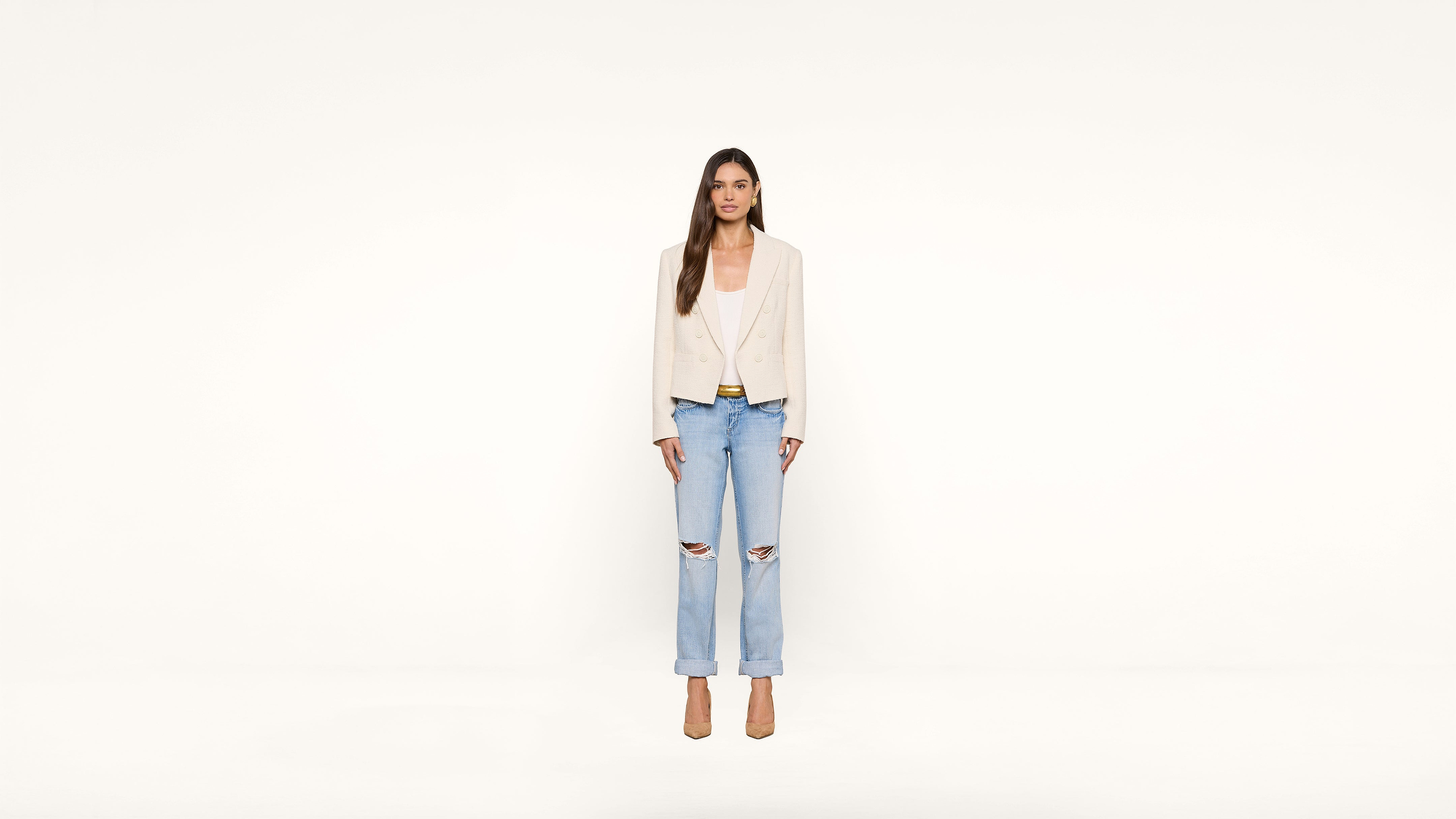 A woman stands against a plain white background, wearing a beige blazer, white top, ripped light blue jeans, and beige heels. She has long brown hair and a neutral expression.