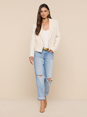 A woman wearing the Brooke Tweed Blazer in ecru, paired with a white top, light blue ripped jeans, and tan heels poses confidently against a beige background. She has long brown hair and one hand in her pocket.