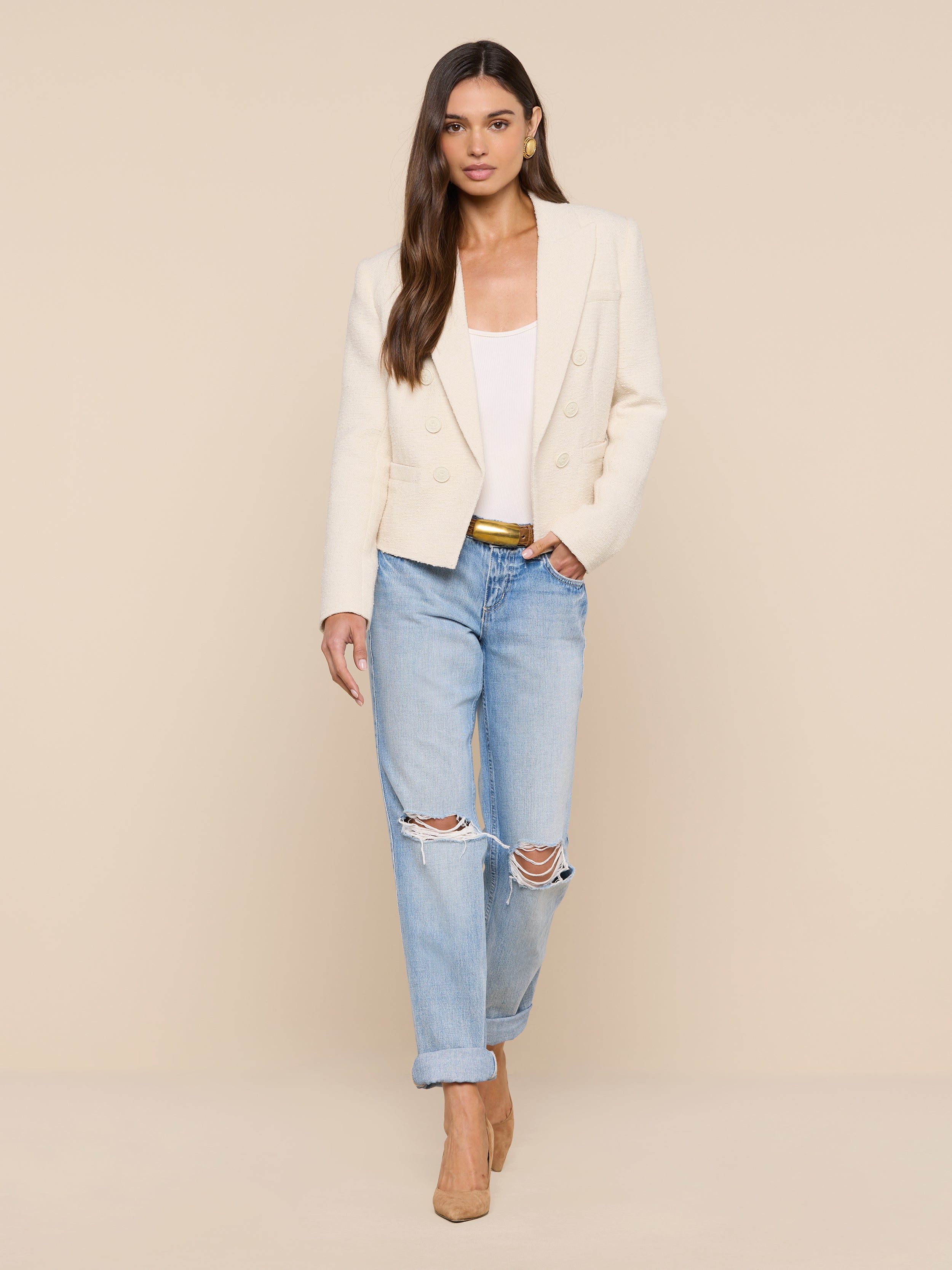A woman stands against a plain white background wearing the Brooke Tweed Blazer over a white top, paired with light blue ripped jeans and beige heels, with one hand in her pocket.