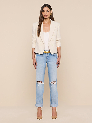 A woman with her hair down and a neutral expression stands against a beige background, wearing the Brooke Tweed Blazer, a white top, light blue ripped jeans, a gold belt, and beige high heels.
