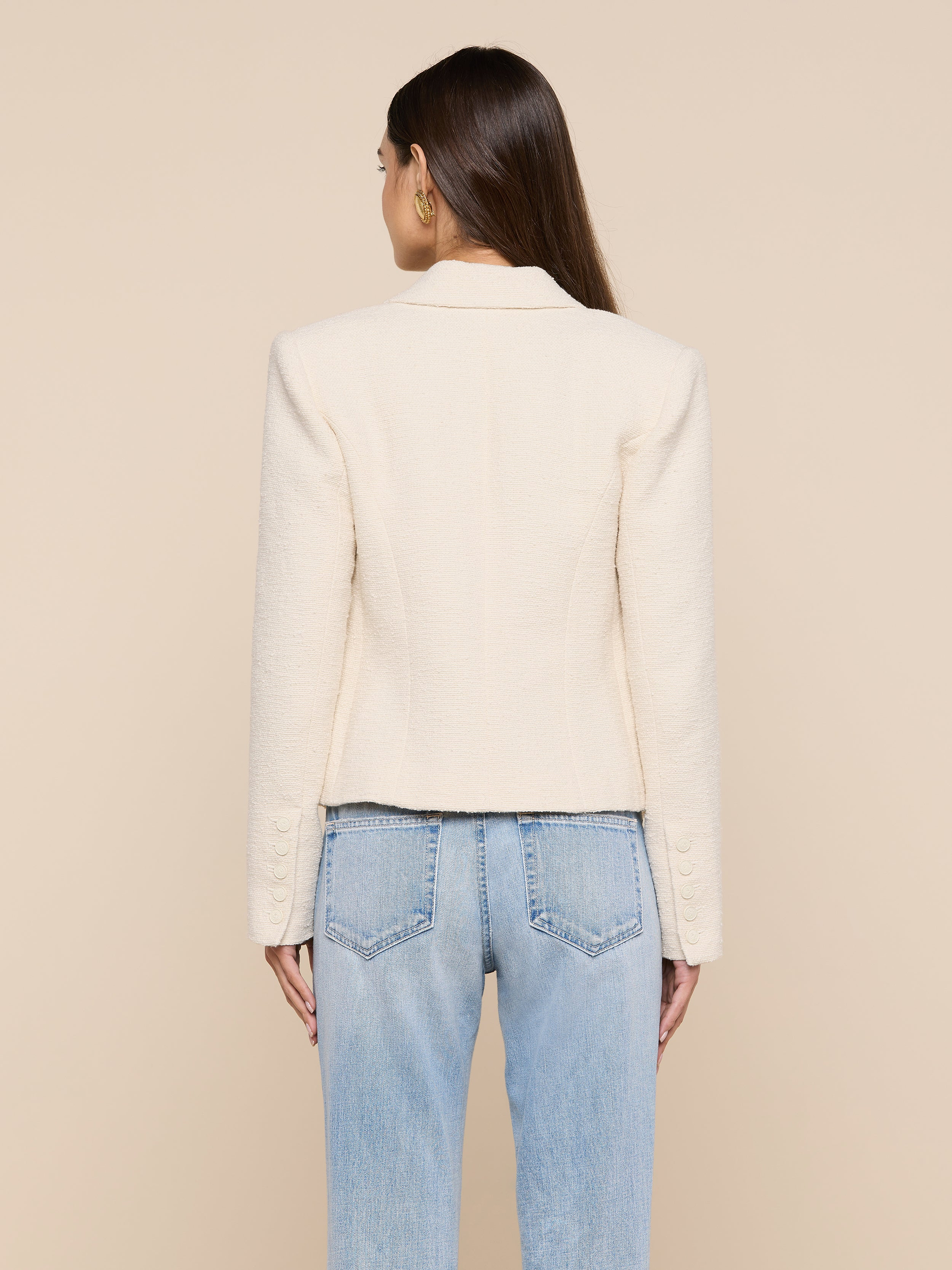 A woman stands facing away, wearing the Brooke Tweed Blazer in a cream hue paired with light blue jeans. Her straight brown hair cascades down her back, set against a plain, light-colored background.