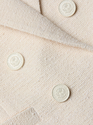 Close-up of Brooke Tweed Blazer’s cream-colored, textured fabric with four embossed cream buttons, each displaying a circular crest and laurel wreath motif. The soft woven tweed showcases elegant detailing.