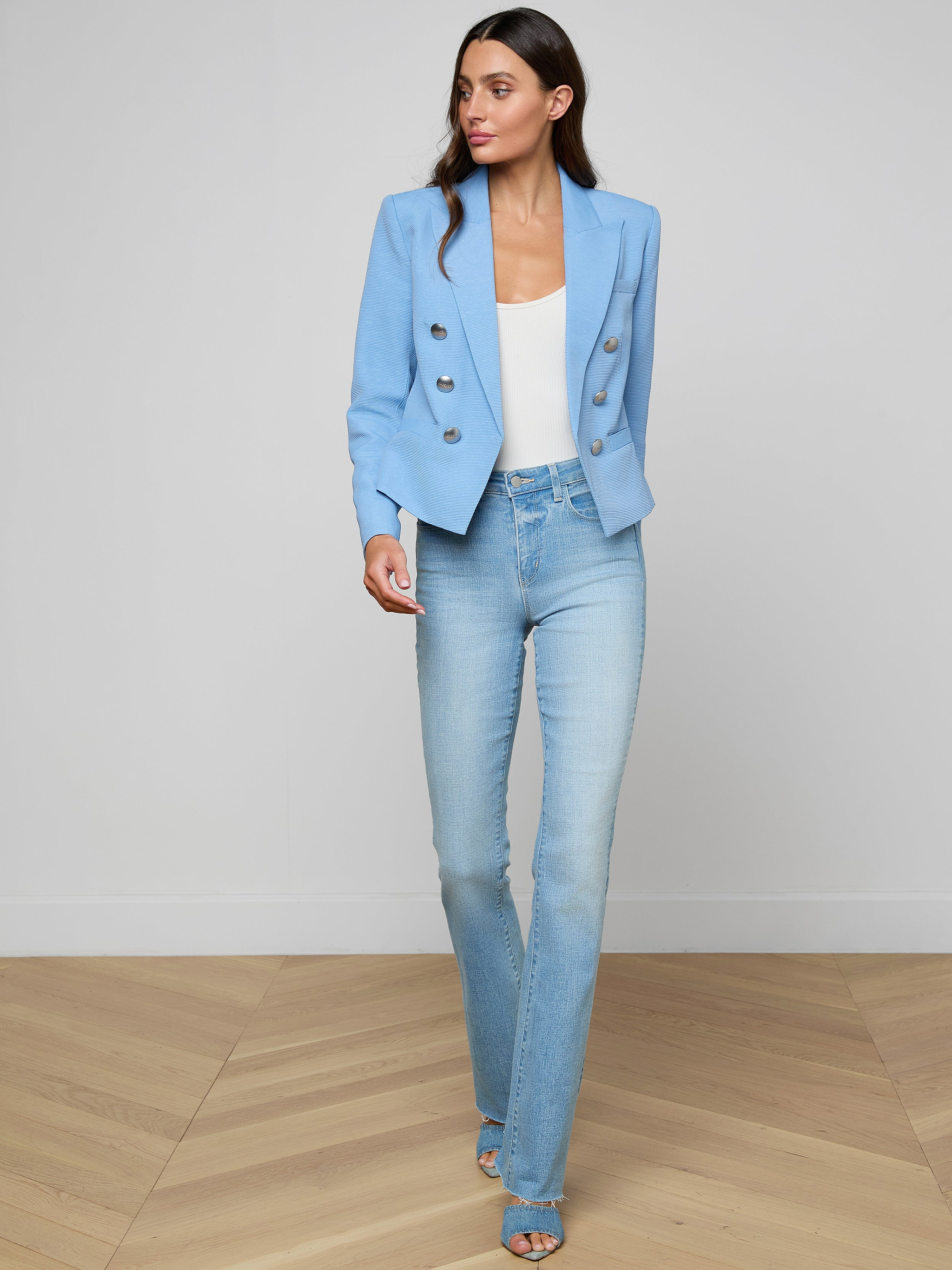 A woman with long dark hair stands against a white background, dressed in the Brooke Blazer—a light blue double-breasted blazer—paired with a white top, light blue jeans, and blue heeled sandals. She is looking slightly to her left.