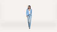 A woman wearing a light blue blazer, white top, blue jeans, and blue heels walks forward against a plain white background.