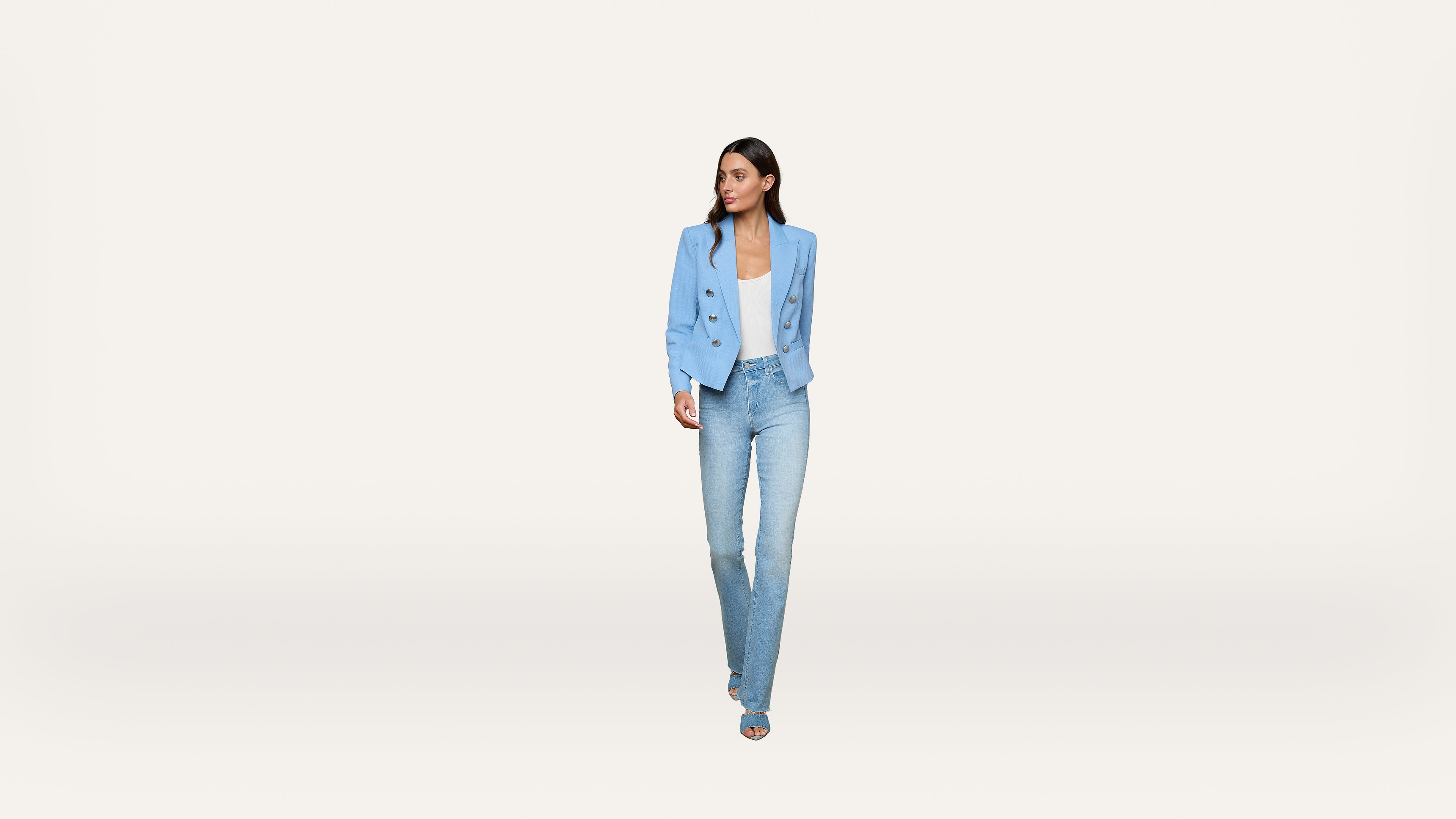 A woman wearing a light blue blazer, white top, blue jeans, and blue heels walks forward against a plain white background.