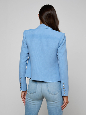 A woman with long brown hair is seen from the back in a Brooke Blazer—a light blue double-breasted style with silver sleeve buttons—paired with matching skinny jeans, against a plain neutral background.