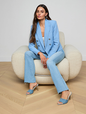 A woman with long brown hair sits on a plush cream armchair, wearing an open-front Brooke Blazer, light blue jeans, and blue heeled sandals. She looks forward with a relaxed expression.