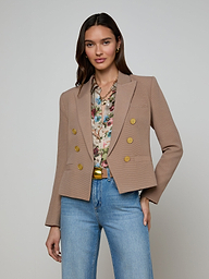 A woman stands against a plain background wearing the Brooke Blazer, an open-front double-breasted style with gold buttons, paired with a floral blouse and blue jeans. Her hand rests in her pocket and she is smiling slightly.